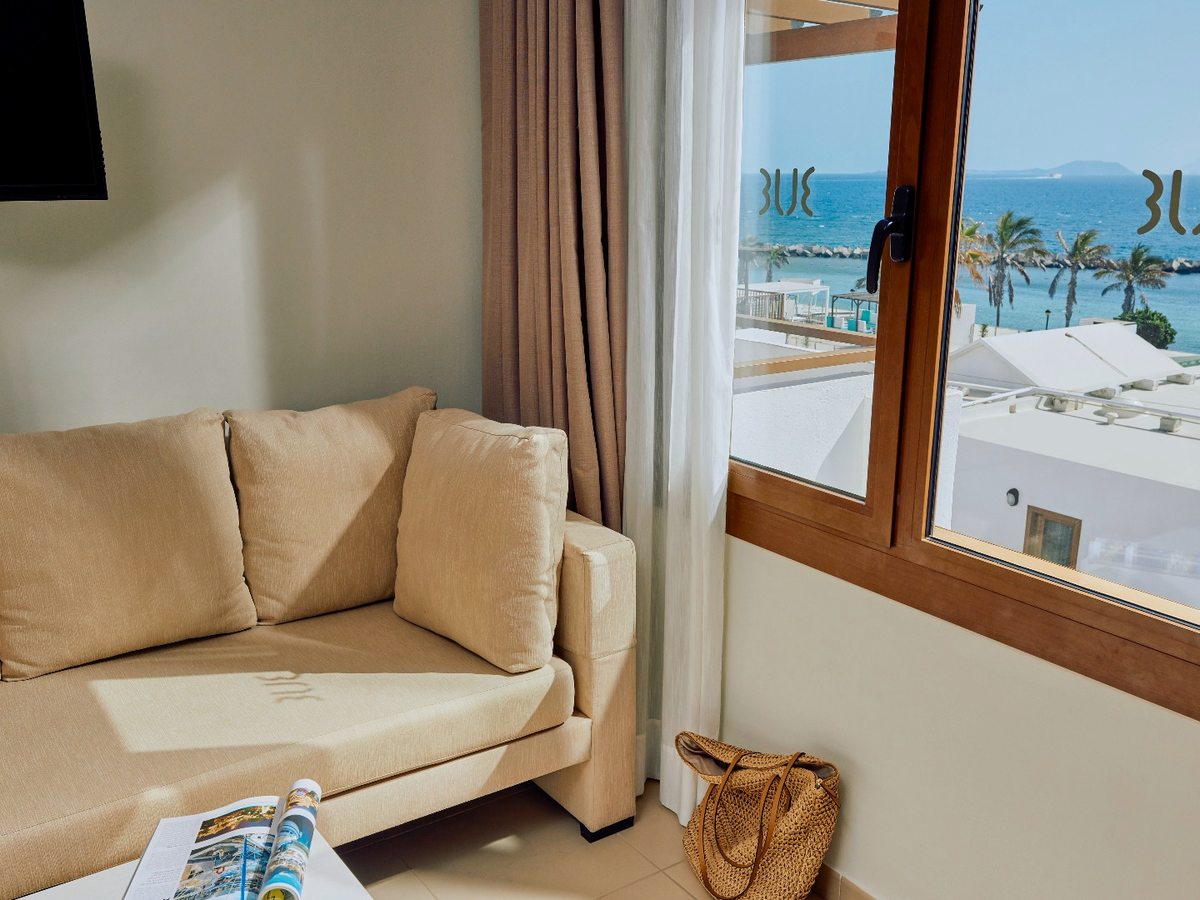 A cream sofa in an apartment lounge beside a wide window showing palms, white resort roofs and the sea, with a wall-mounted television to the left.