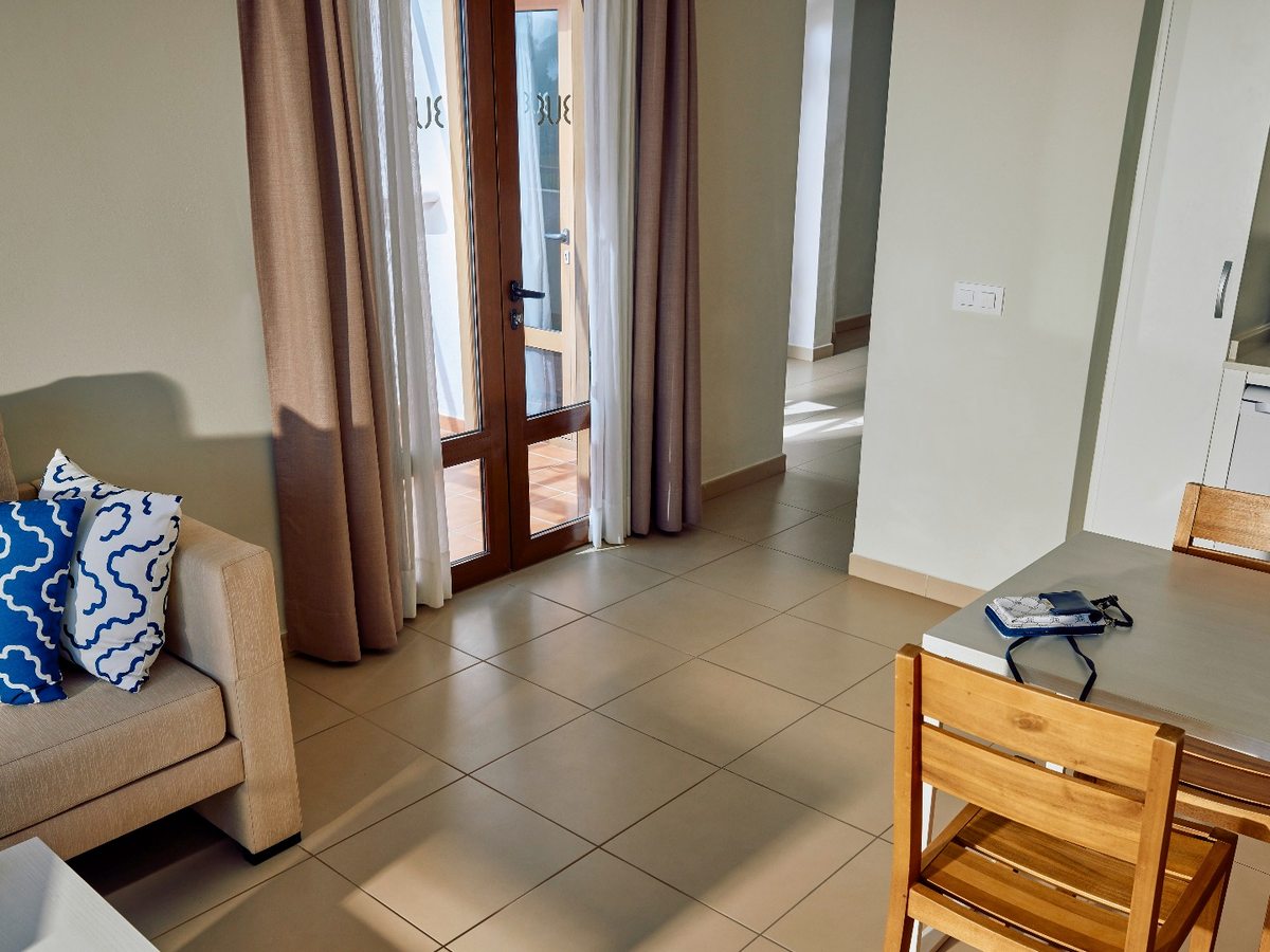An open-plan apartment living area with a beige sofa, a small wooden dining table and chairs, a tiled floor and glazed doors opening onto a terrace.