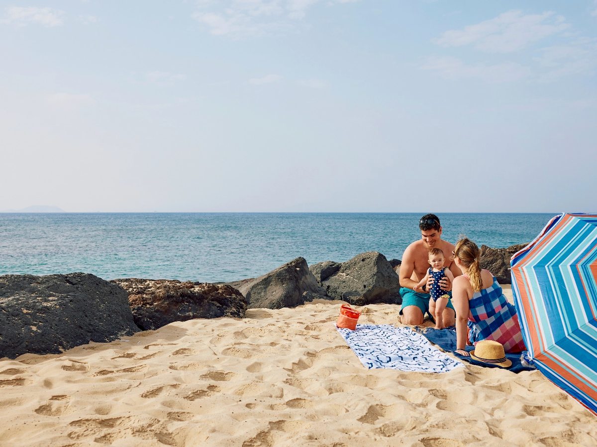 A family with a baby sitting on soft golden sand beside dark volcanic rocks under a striped umbrella, with calm blue sea behind.