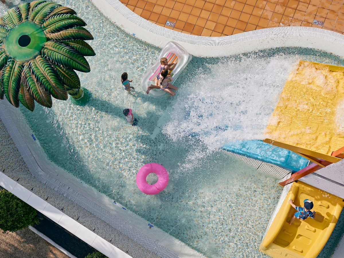 An overhead view of a shallow splash pool with children playing, a large green palm-tree water feature spraying water and yellow and blue slides running into the water.