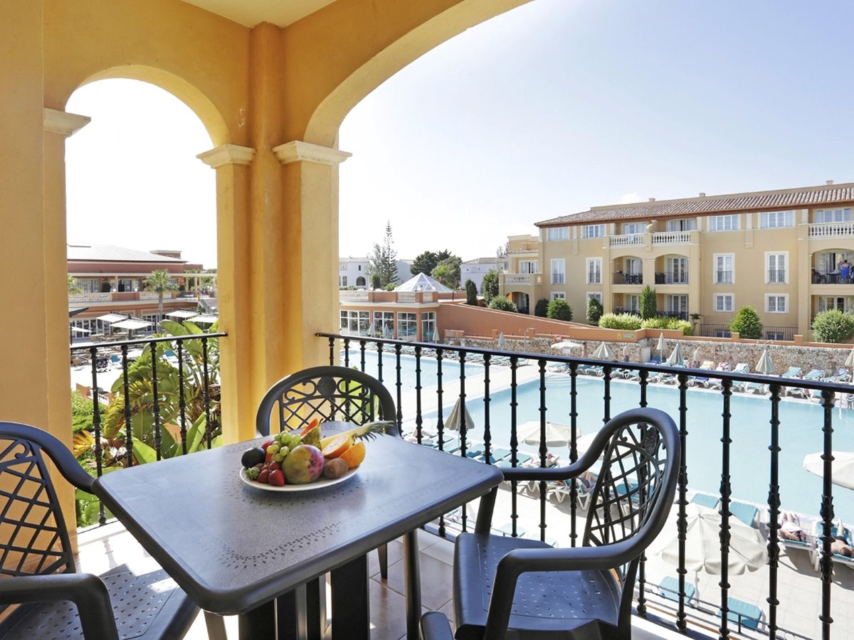A shaded terrace behind yellow arches with a table, a bowl of fruit and dark metal chairs, looking down over the main pool, its loungers and the apartment blocks beyond.