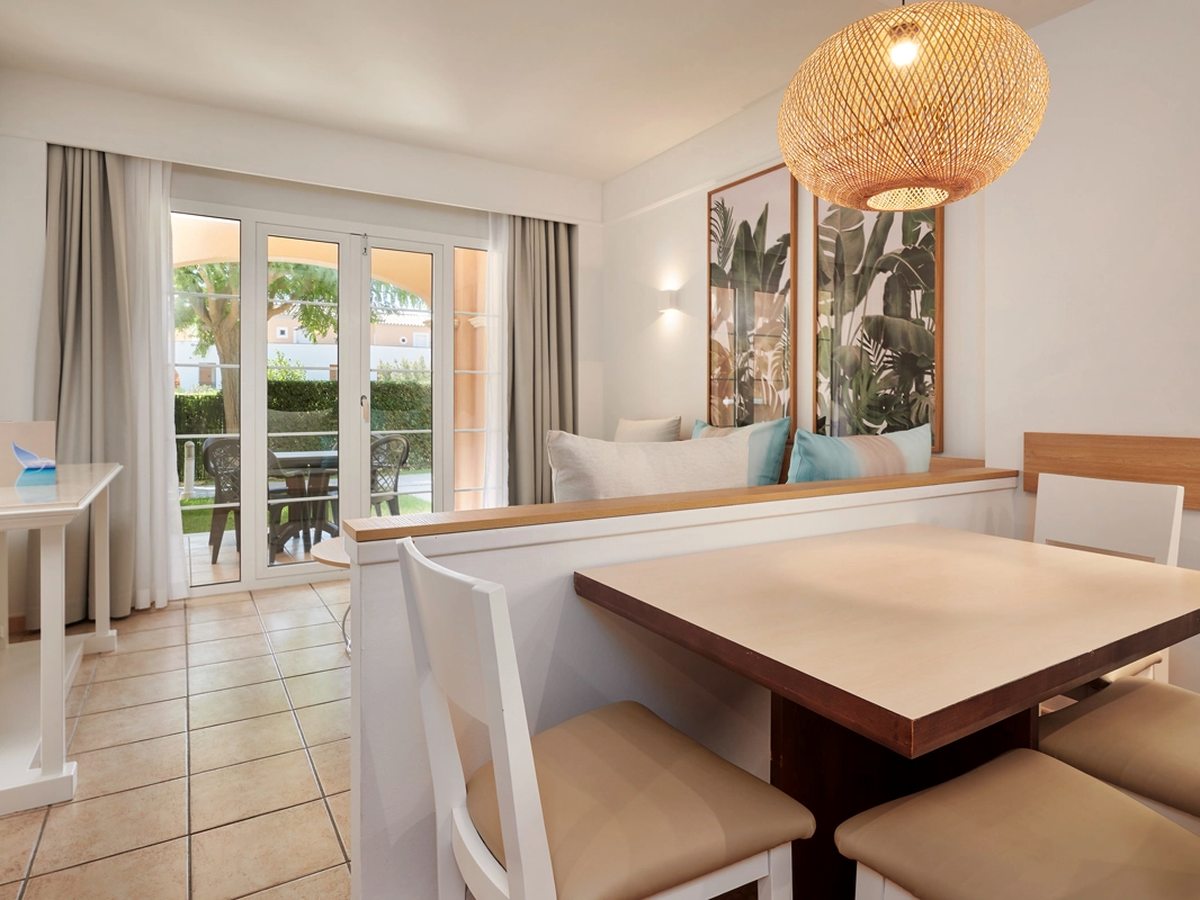 A four-seat dining table under a woven pendant light, with a half-height divider between it and the sofa in the living area and terrace doors beyond.