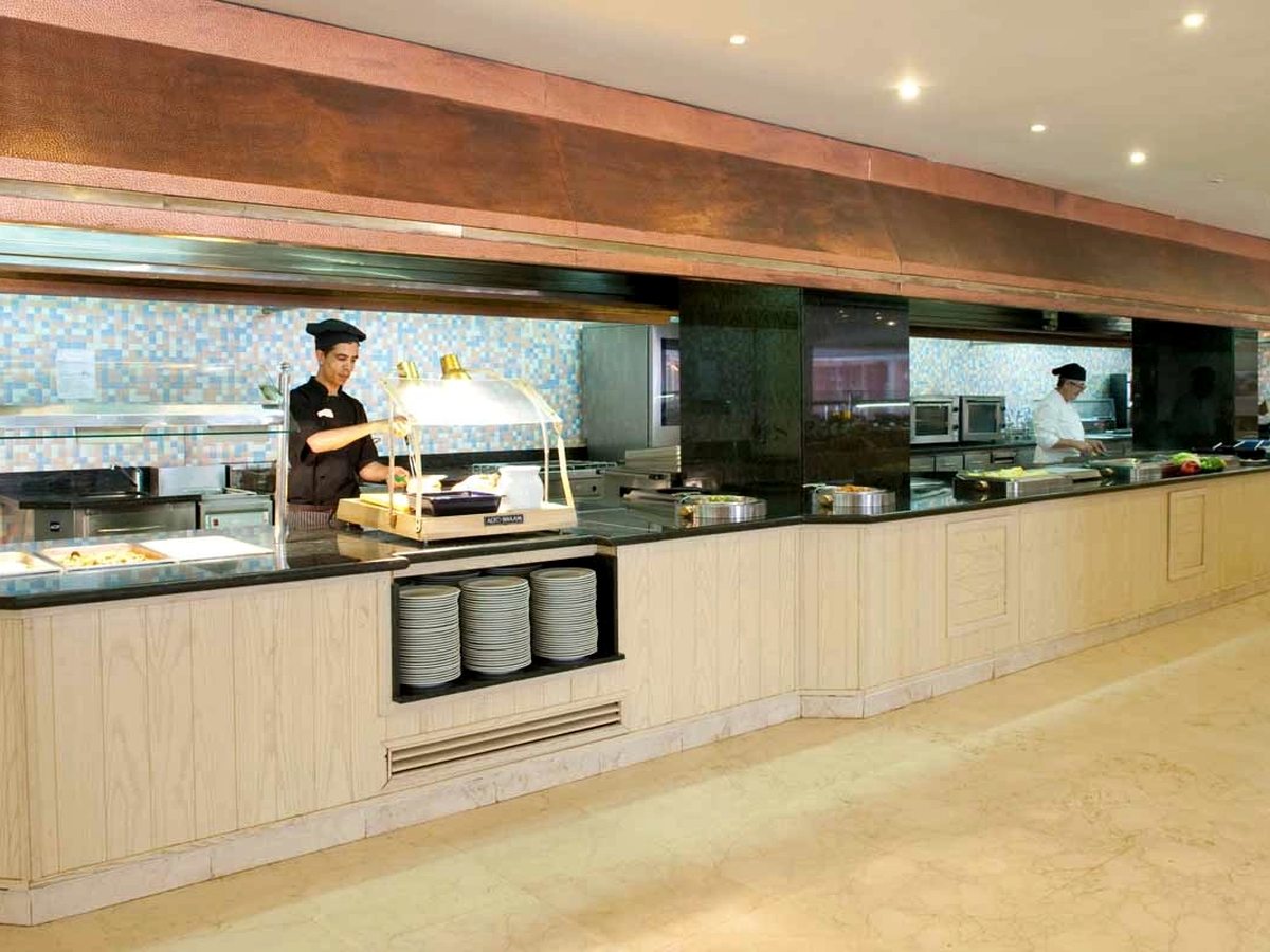 A chef in whites standing behind a hot buffet counter with stacked plates in front of him and an open kitchen behind.