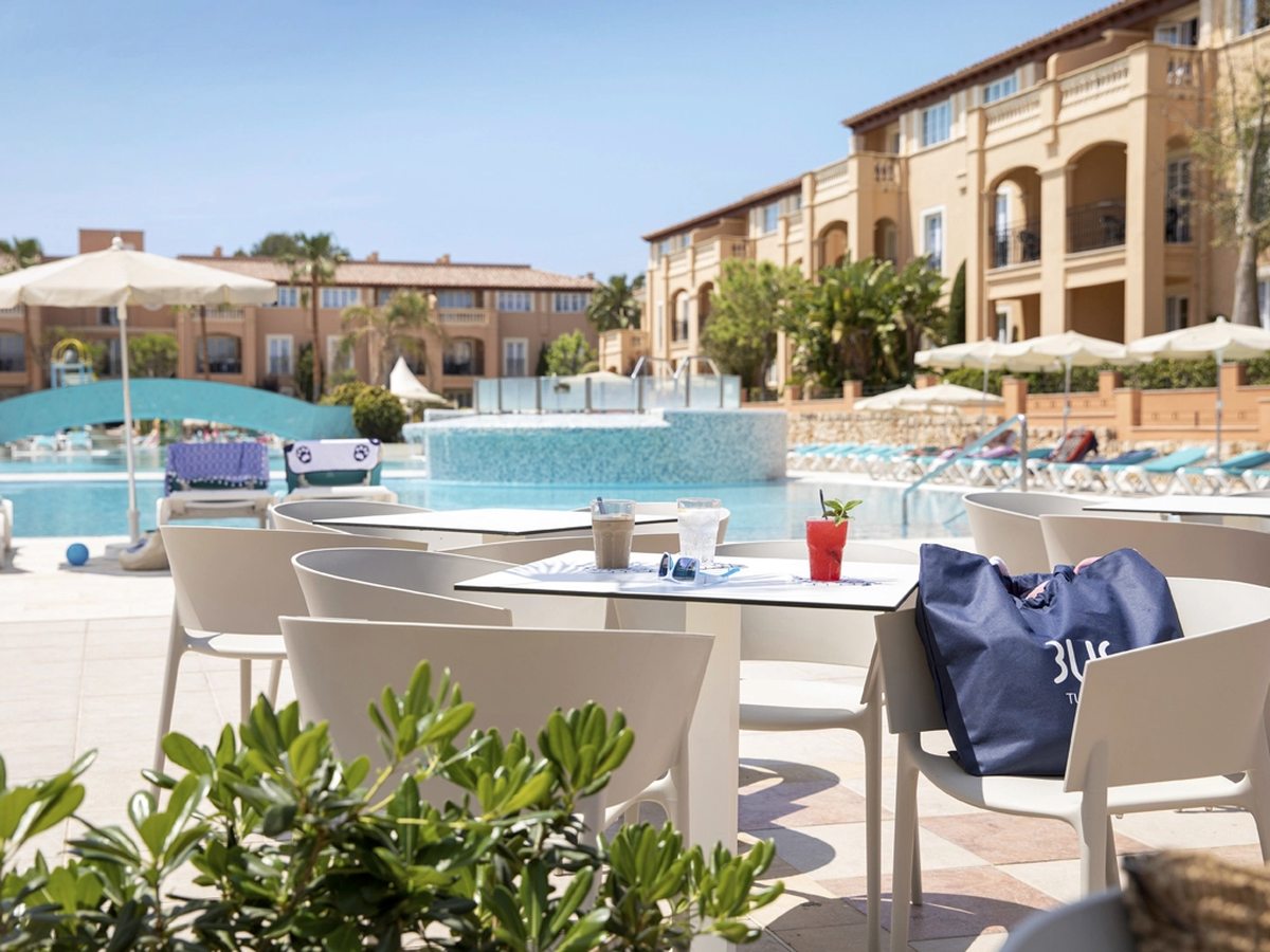 White tables and chairs on a paved terrace beside the main pool, drinks on the table in front and the apartment blocks and parasols behind.