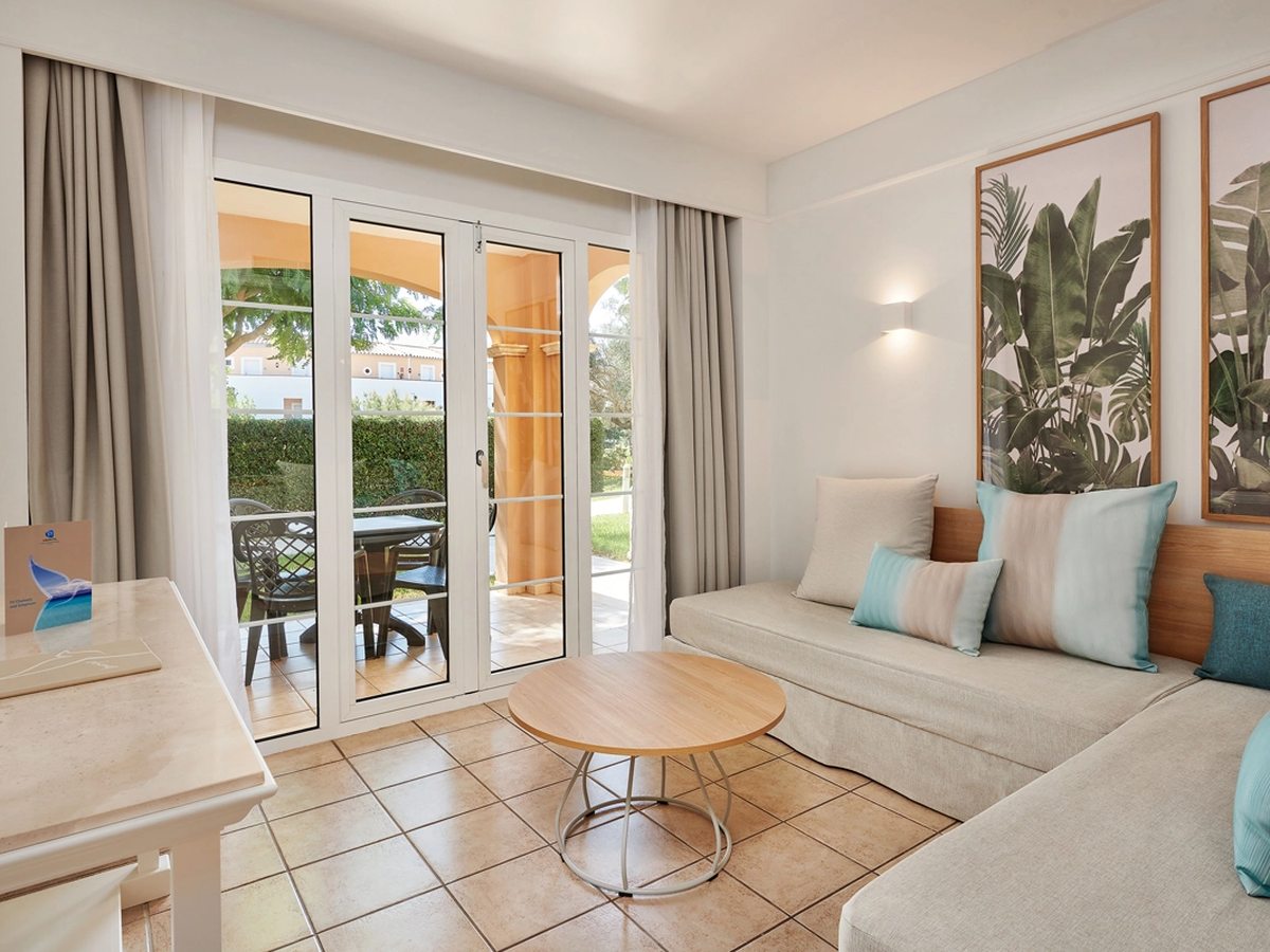 An apartment living room with a cream corner sofa, a round wooden coffee table and botanical prints, and glazed double doors to a terrace with a table and chairs beyond.