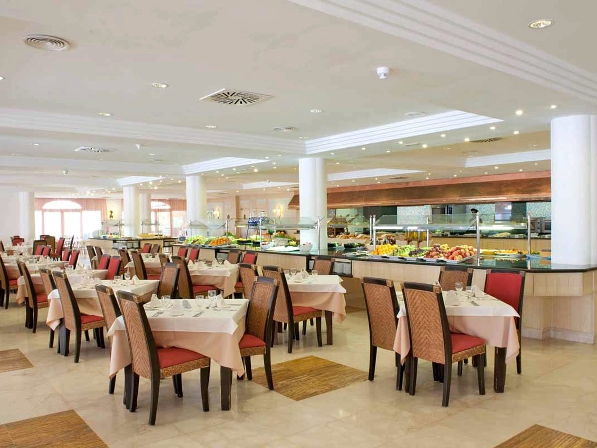 A large buffet restaurant with long serving counters of fruit and salad along one side, white columns and set tables with pink cloths and red chairs.