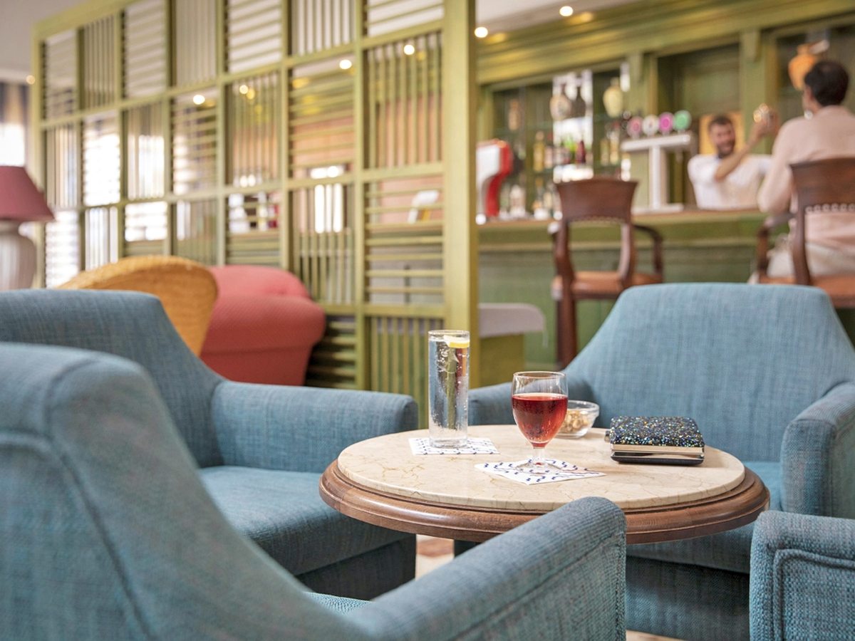 An indoor bar lounge with blue upholstered armchairs around a small round drinks table, a bar counter and panelled green screens behind.