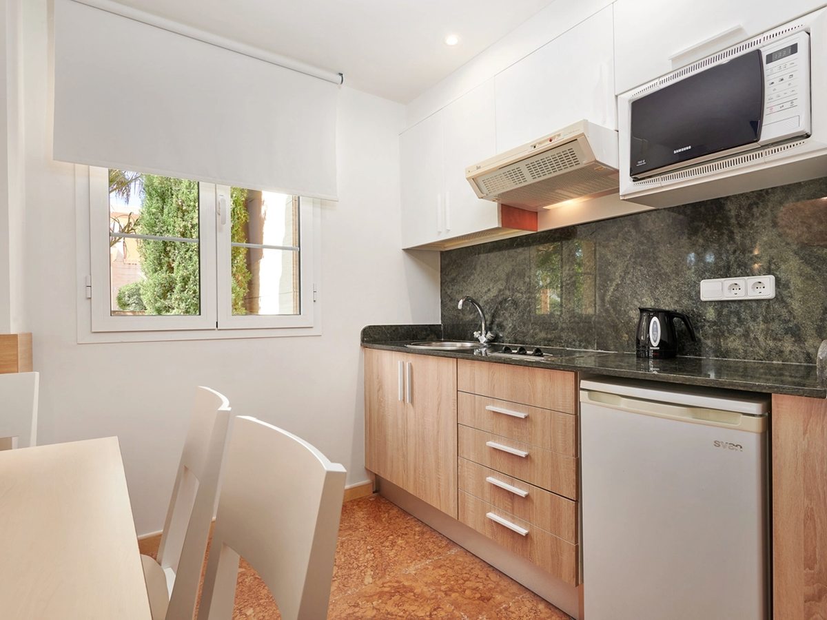 A compact kitchenette with wood-fronted units, a dark stone worktop and splashback, sink, two-ring hob, kettle, microwave above and an under-counter fridge, beside a small dining table.