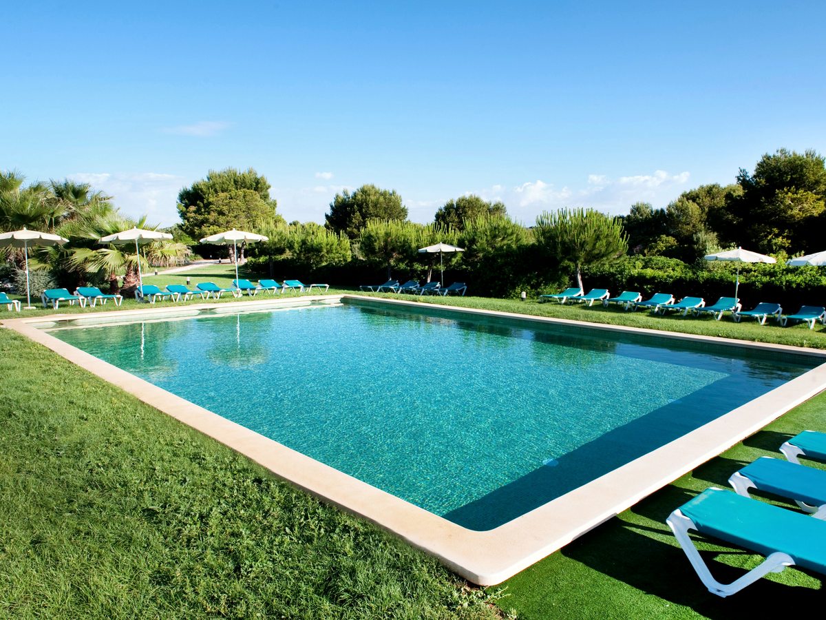 A rectangular pool edged in pale stone and surrounded by mown grass, with turquoise loungers, white parasols, palms and pines behind under a clear sky.