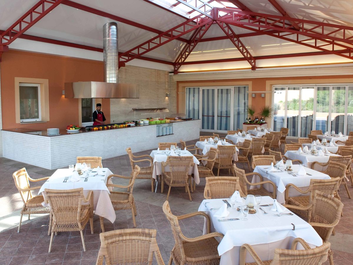 A covered terrace restaurant under a red steel-truss roof, with wicker armchairs at white-clothed tables and a chef at an open cooking counter along one wall.