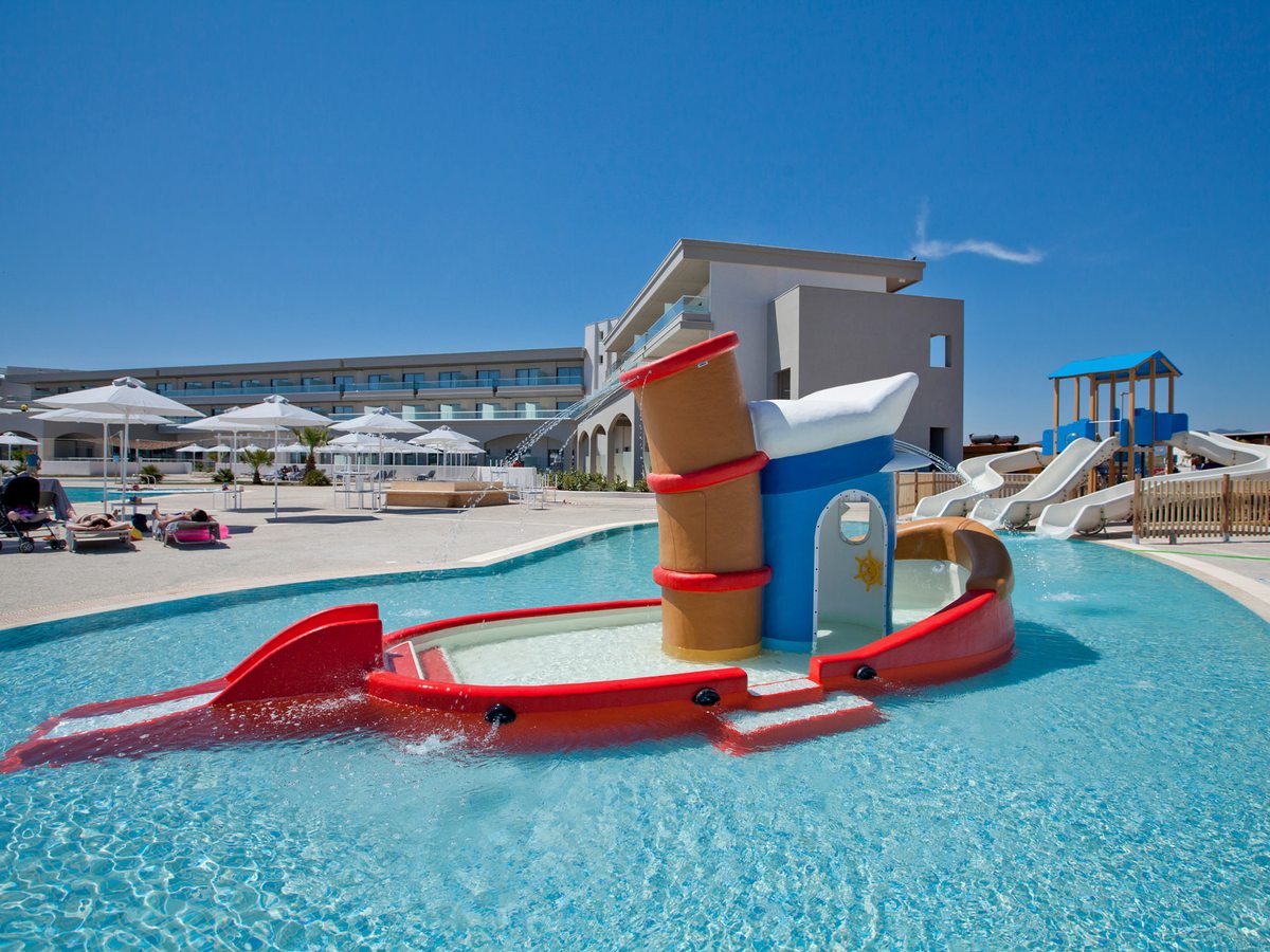 A red and blue tow boat play feature spraying water in a shallow pool, with small white water slides and a play frame behind.