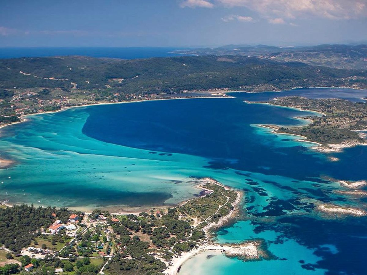 An aerial view of a wide turquoise bay with sandy shallows, a wooded headland and villages scattered along the shore.