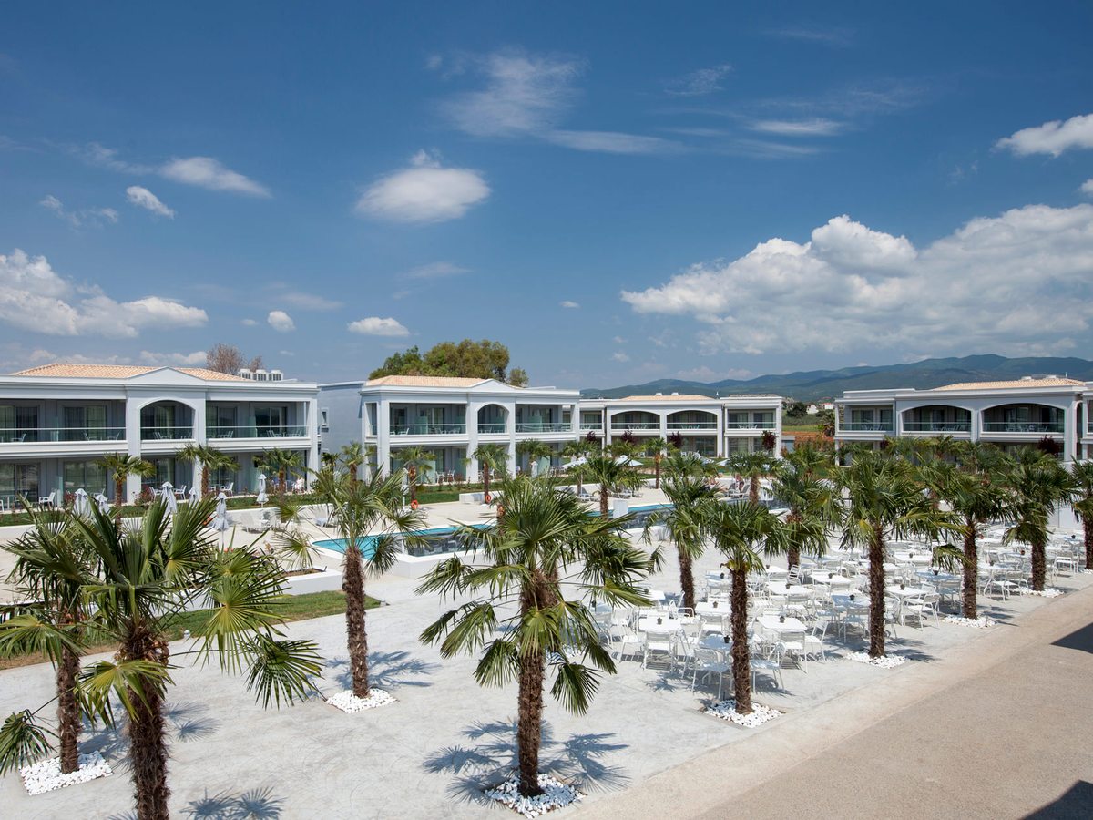 Two-storey guest buildings around a wide paved square planted with fan palms, rows of white tables and chairs to one side and a long narrow pool beyond.