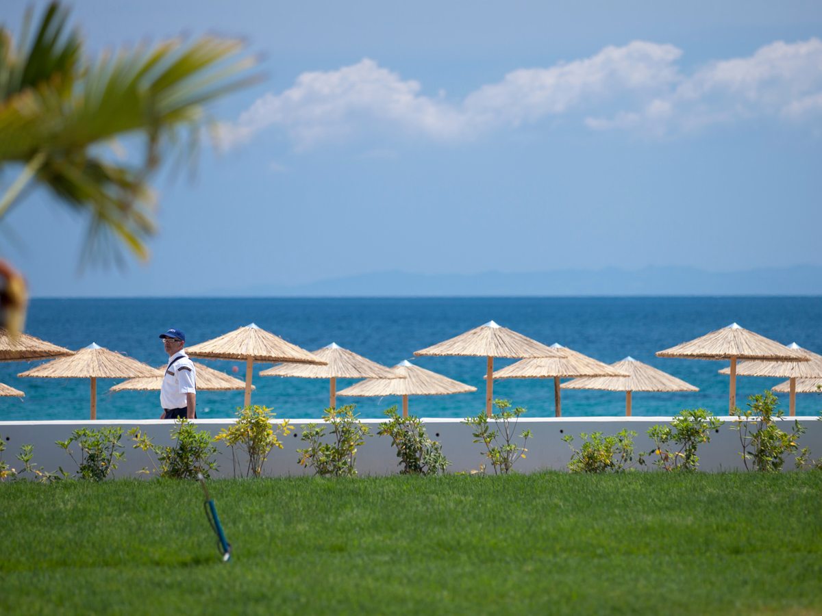A mown lawn running down to a low wall, with a row of straw parasols on the sand beyond and the sea past them.