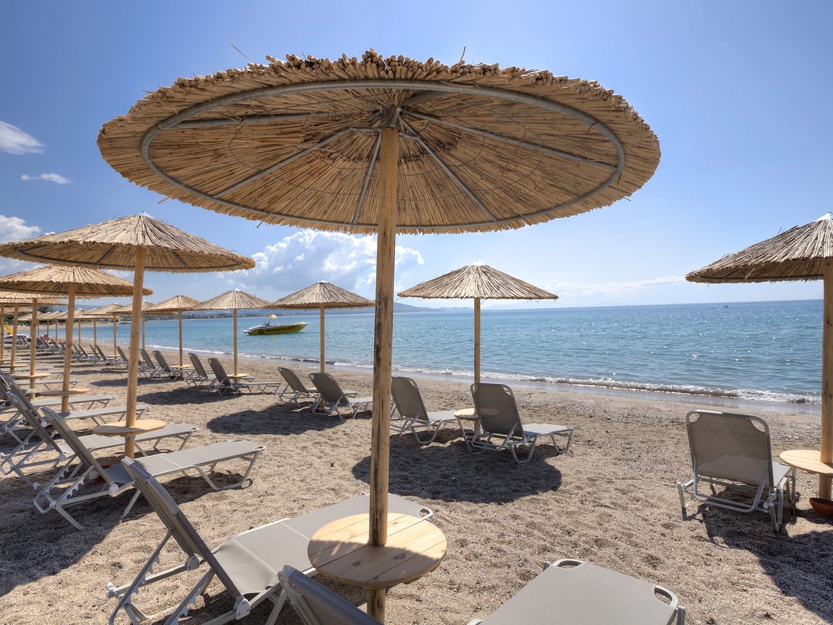 Straw parasols and sun loungers on a beach of coarse sand and pebbles, with calm blue water and hills across the bay.