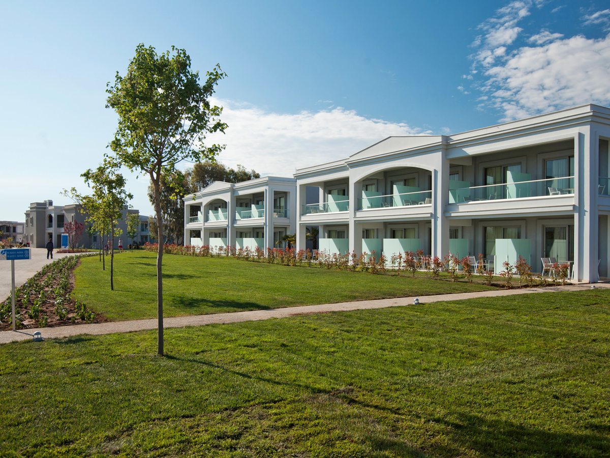 A white two-storey guest building with arched balconies and pale green glass screens, seen across a mown lawn with a young tree and a paved path.