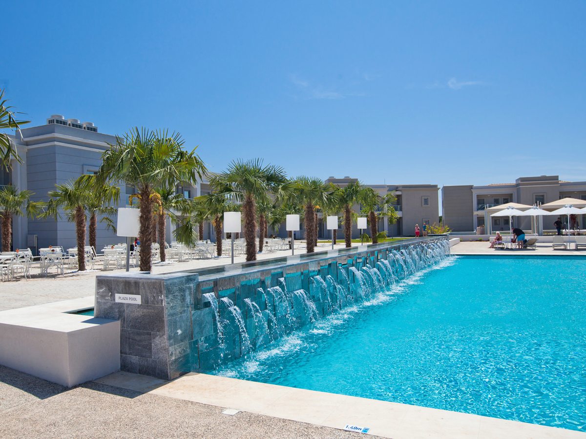 A long pool fed by a row of small waterfalls from a stone wall, with fan palms and white tables and chairs along the terrace above.