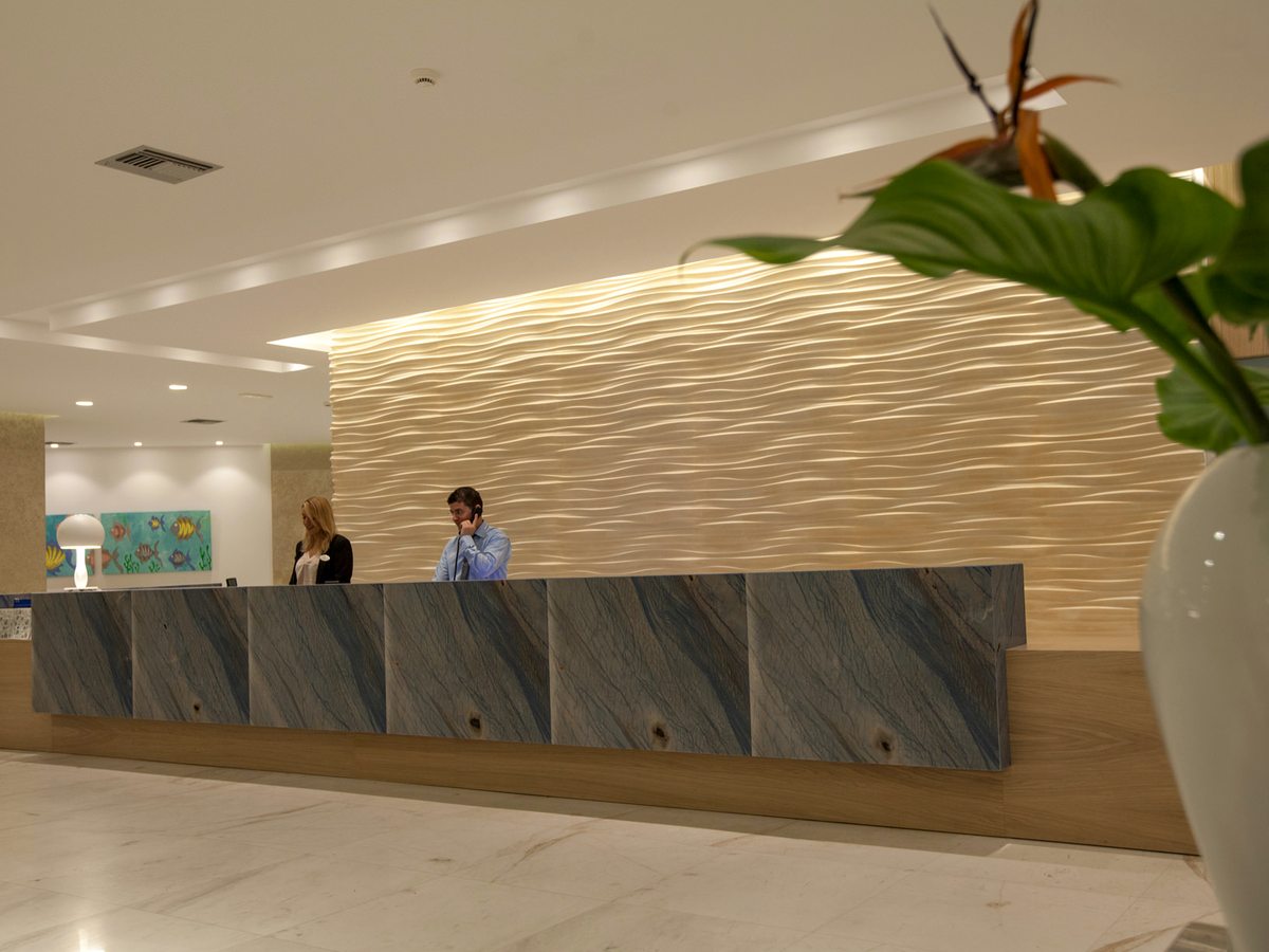 A long reception counter against a textured timber wall, staff behind it, with a polished marble floor and a potted plant in the foreground.