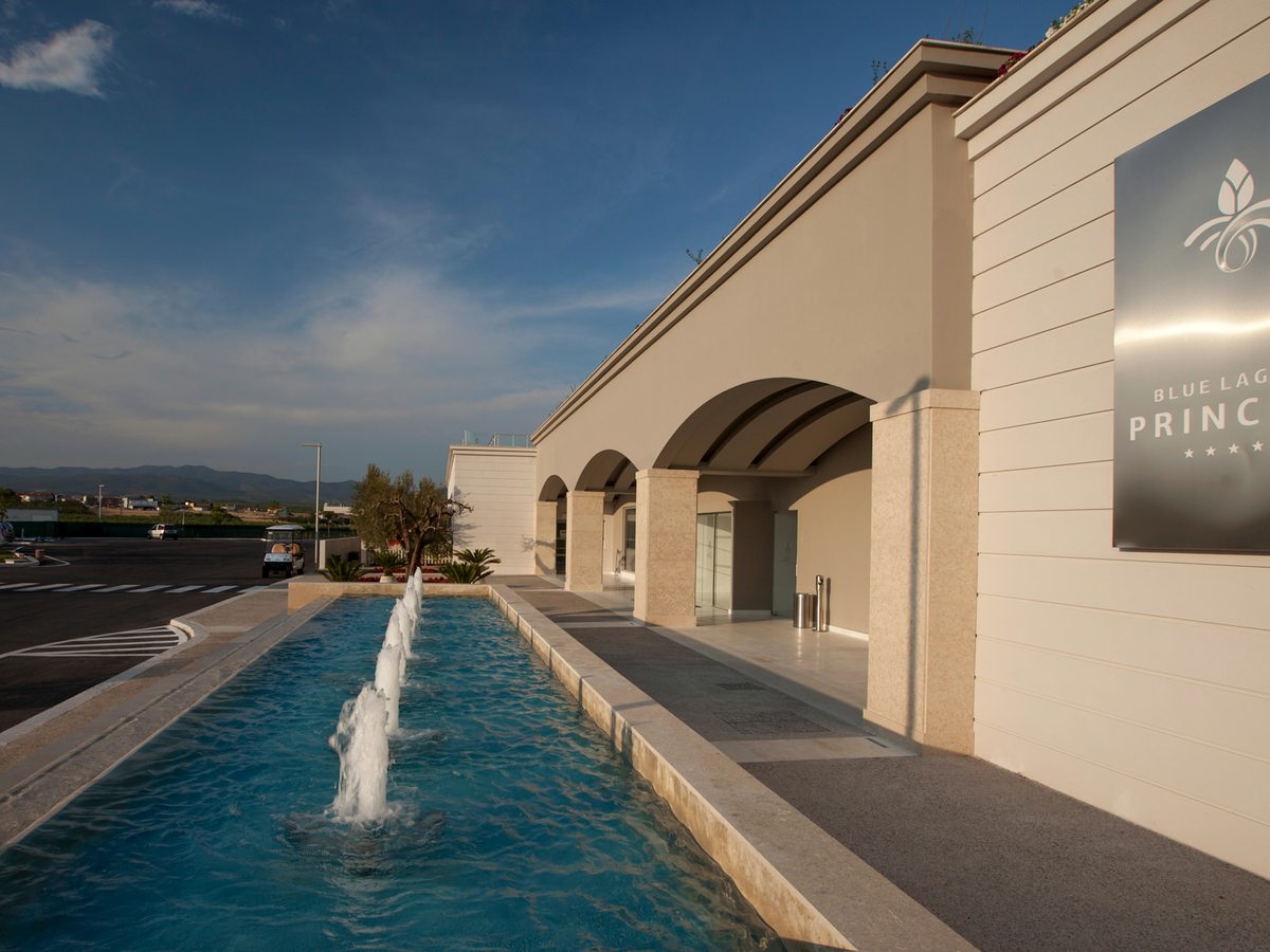 The arcaded front of the main building beside a narrow water feature with fountain jets, the Blue Lagoon Princess sign on the wall and a golf buggy parked on the drive.