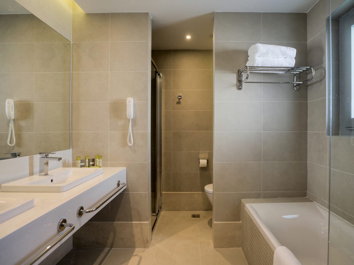 A bathroom with twin basins on a long counter and a mirror above, a glazed shower cubicle, a toilet and a bath with folded towels.