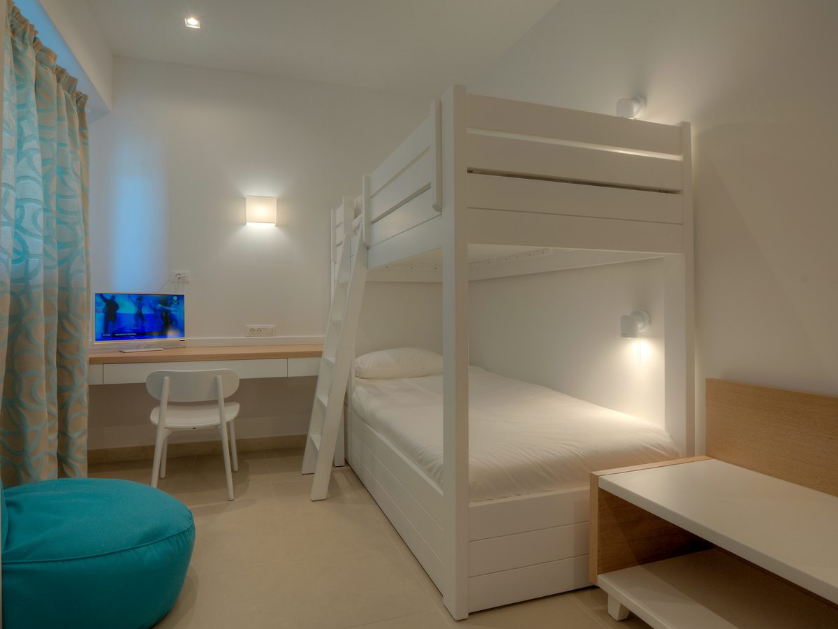 A children's bedroom with white wooden bunk beds and a ladder, a built-in desk with a small television and a turquoise floor cushion.