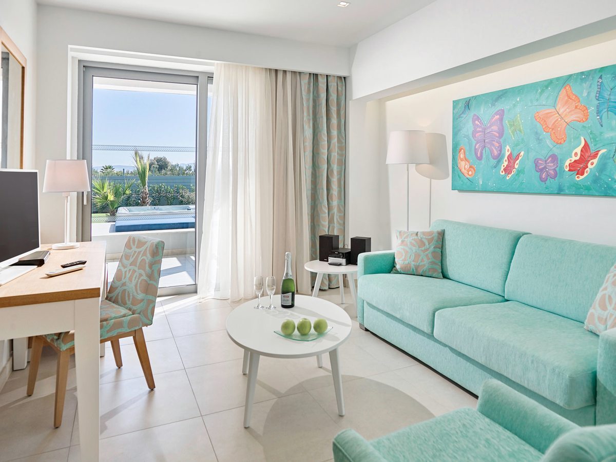 A suite living area with a turquoise sofa and armchair, a round coffee table, a desk and butterfly artwork, with terrace doors onto a pool.