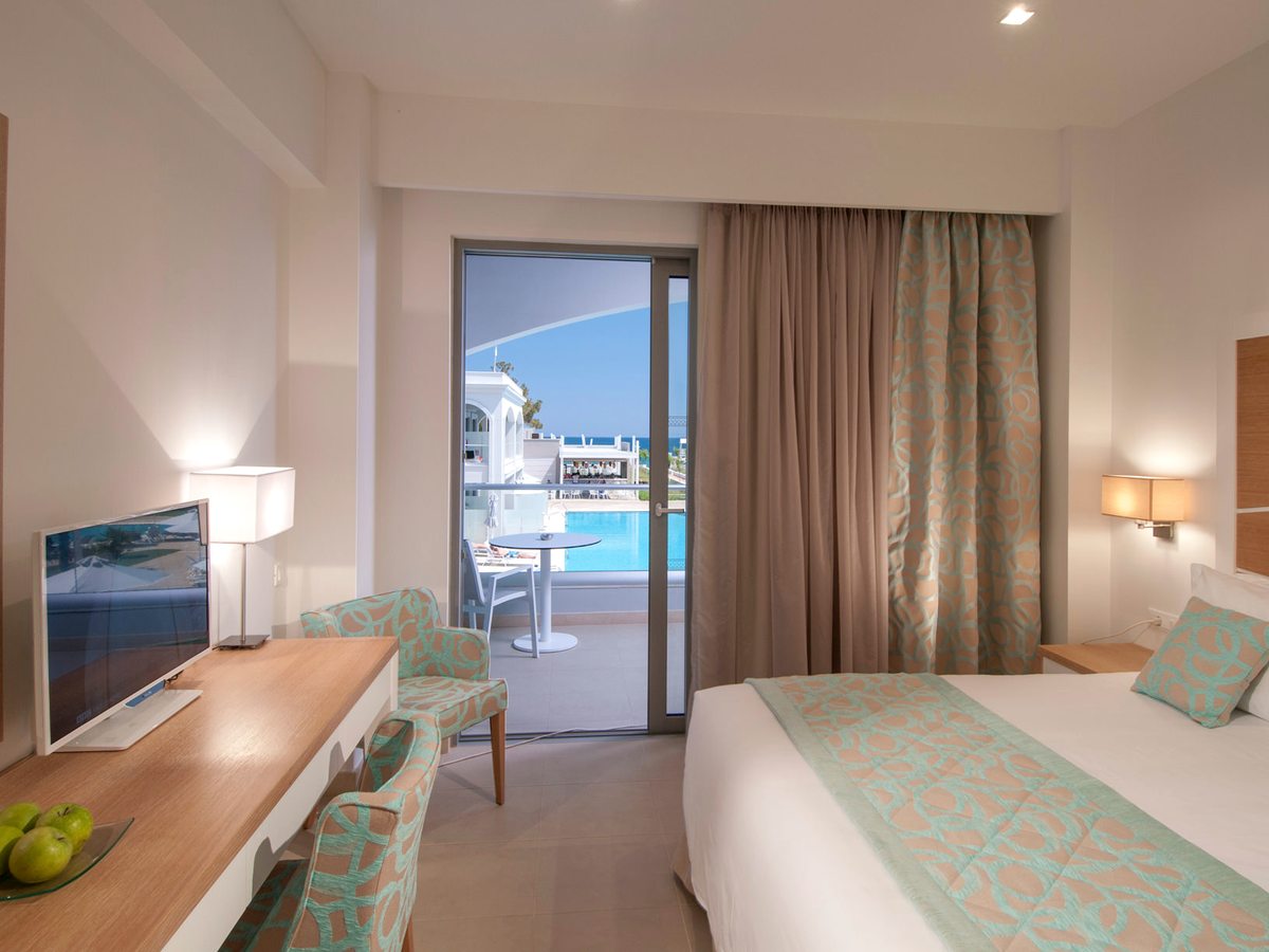 A room with a bed, desk and television, its balcony doors open to a small table and chairs above a blue pool.
