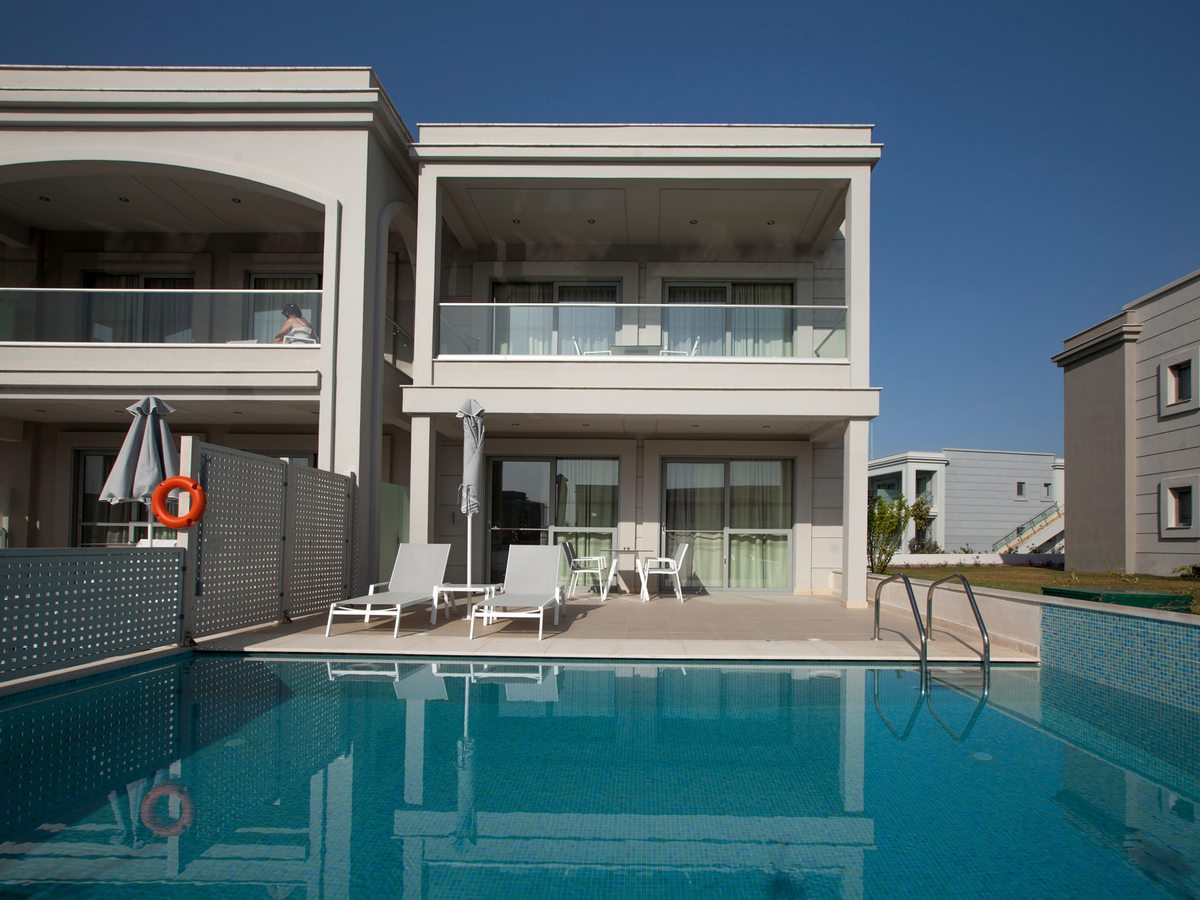 A pool running up to the tiled terraces of ground-floor rooms, with sun loungers, a folded parasol and metal steps into the water.