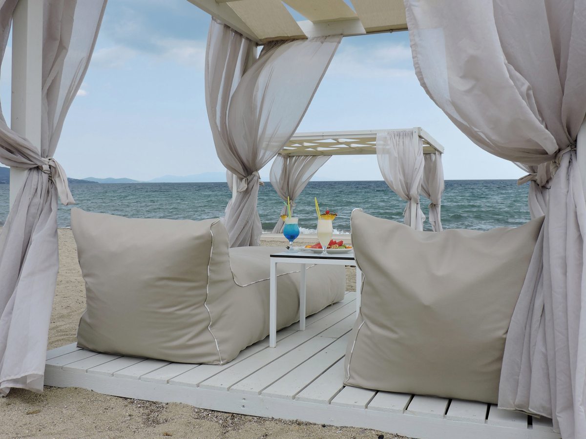 A white curtained daybed on the sand piled with cushions, a small drinks table beside it and the sea beyond.