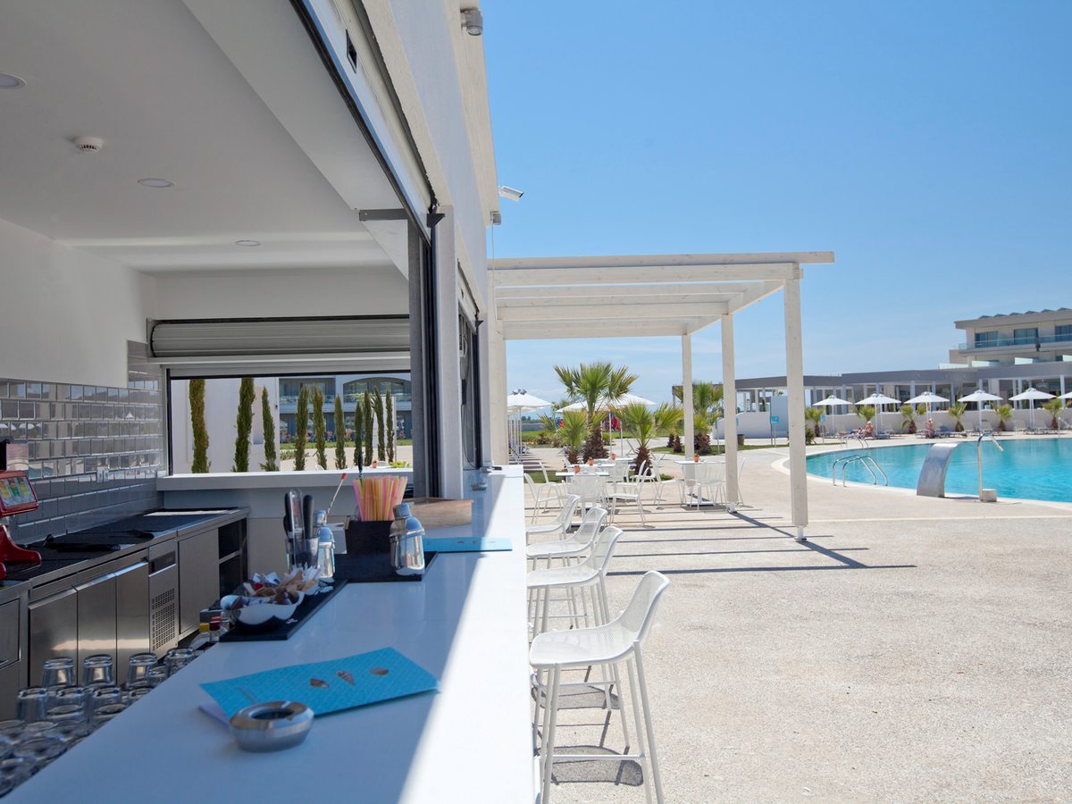 A poolside bar with a white counter, glasses and bar stools, a shaded pergola with tables alongside and the blue pool beyond.