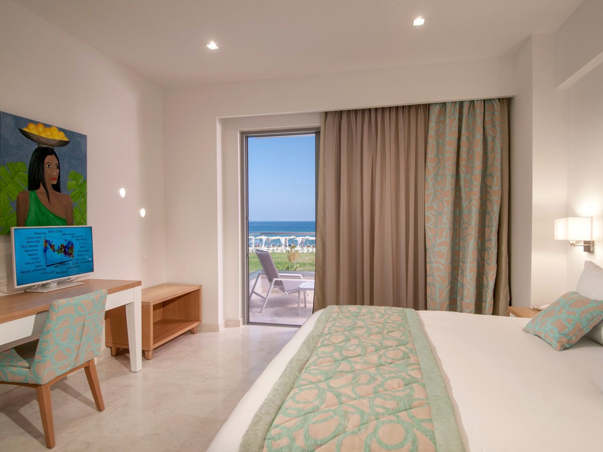 A room with a large bed, a desk and television, and open patio doors to a balcony with chairs and a view over beach parasols to the sea.