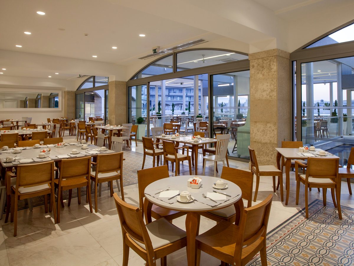 An indoor restaurant with round and square tables and wooden chairs, arched glass partitions along one side opening to a terrace with more tables.