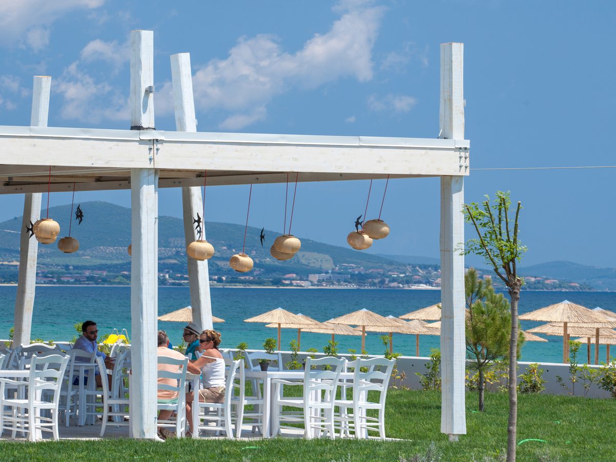 White tables and chairs under a timber pergola hung with woven lanterns, on a lawn above a beach lined with straw parasols and calm blue sea.