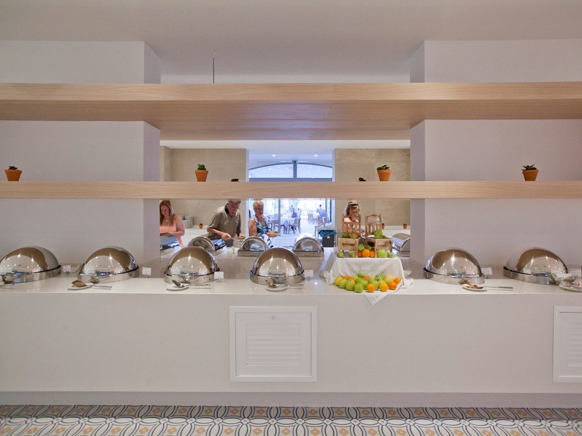 A long white buffet counter set with domed silver chafing dishes and a crate display of apples and oranges, guests serving themselves from the far side.