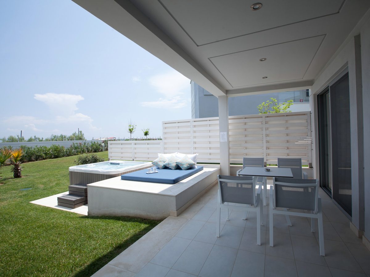 A covered room terrace with a dining table and chairs and a cushioned daybed, and a free-standing jacuzzi with steps up to it on the paving alongside.