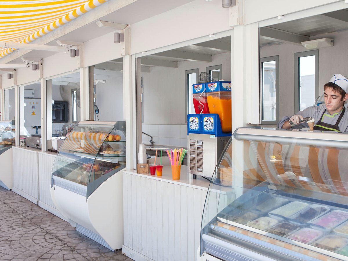 A snack kiosk with ice cream and pastry display cabinets and slush machines, a member of staff scooping a cone at the counter.