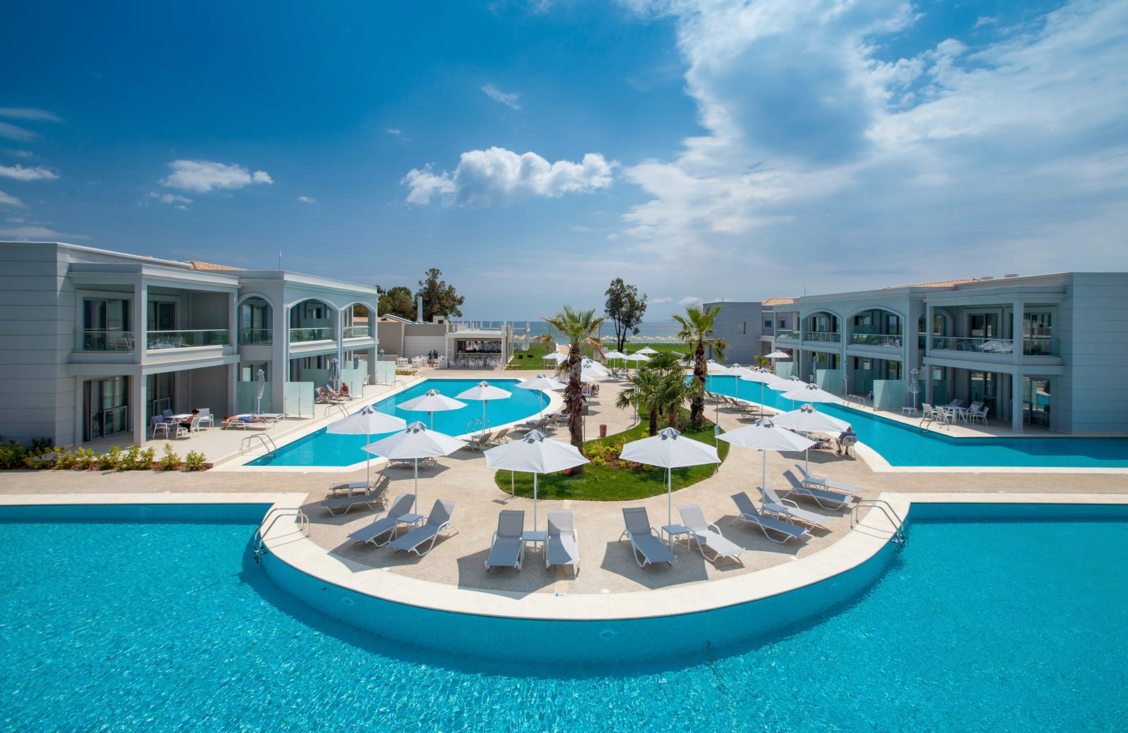 Pools winding between the low-rise white buildings of TUI BLUE Lagoon Princess, with sun loungers and parasols on the terraces between them and the sea beyond.