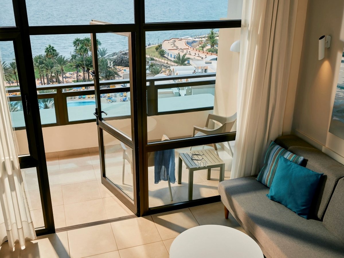 A tiled room with a grey sofa and a round white table, its sliding doors open onto a balcony above the pool deck, the palm-lined promenade and the sea.