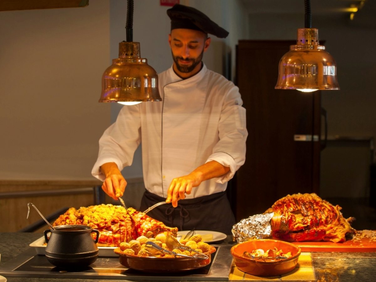A chef in whites and a black hat serving roast meat and potatoes from trays under two copper heat lamps at a buffet counter.
