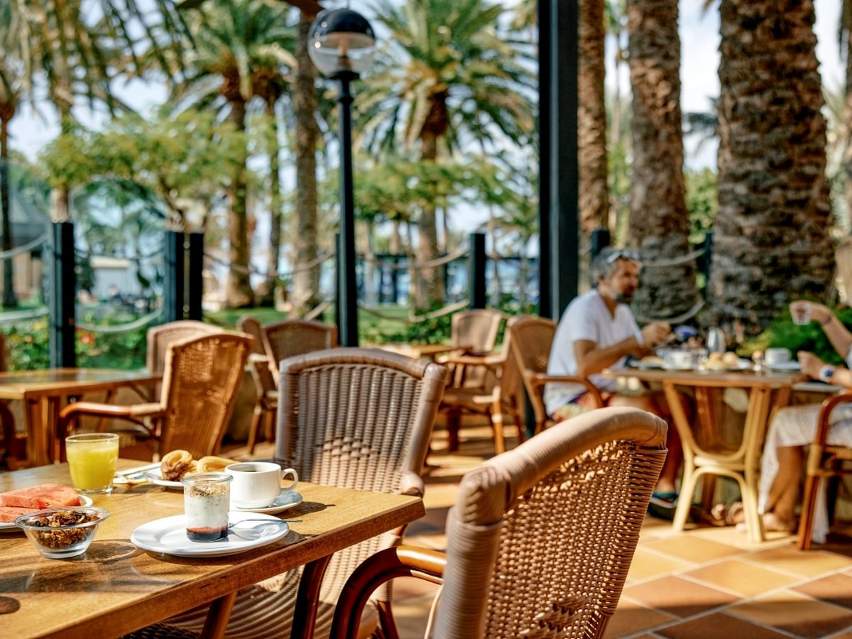 A shaded terrace with wicker chairs and wooden tables set with coffee, orange juice, watermelon, granola and pastries, with tall palms and a lawn beyond.
