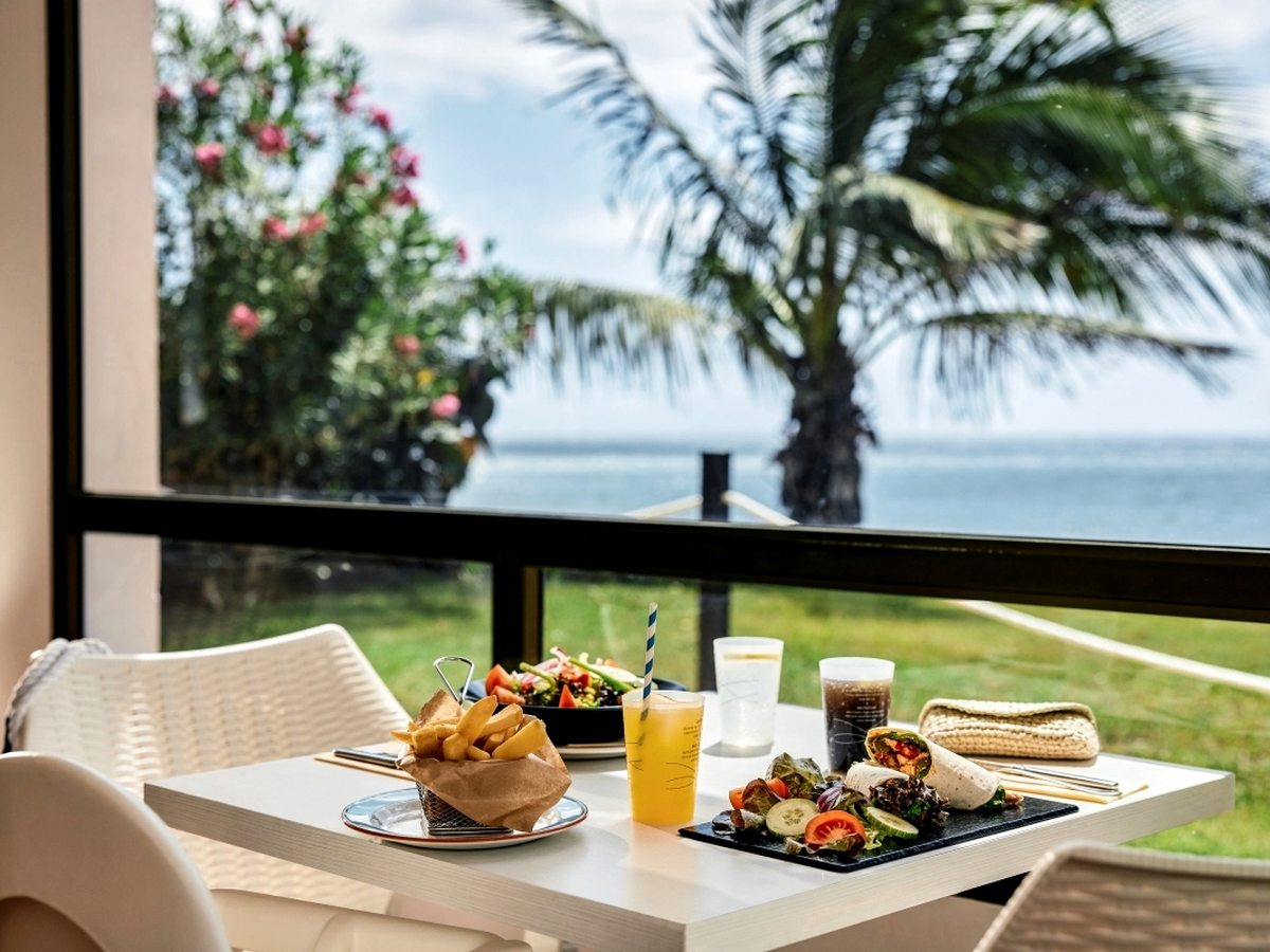 A white table set with a wrap, a salad and a basket of chips beside cold drinks, in front of a window onto palms, pink oleander and the sea.