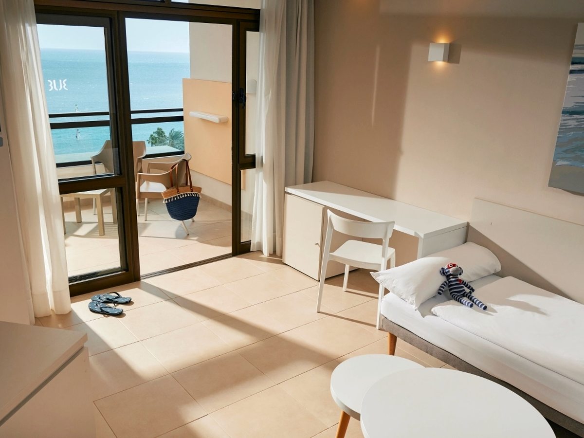 A sunlit tiled room with a single bed made up along the wall, a white desk and chair, and open sliding doors onto a balcony above the sea.
