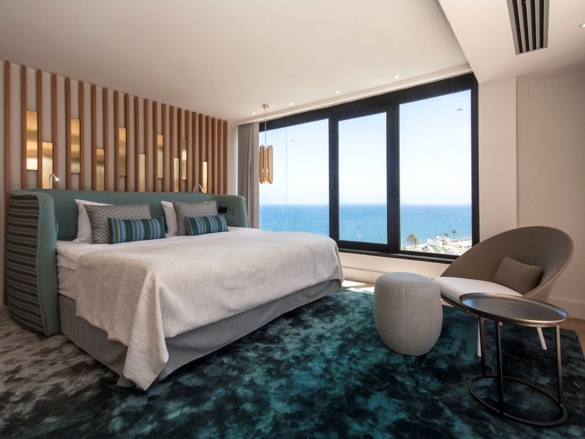 A bedroom with a wide bed against a slatted timber and brass wall, a deep teal carpet, a round armchair and a window onto the sea.