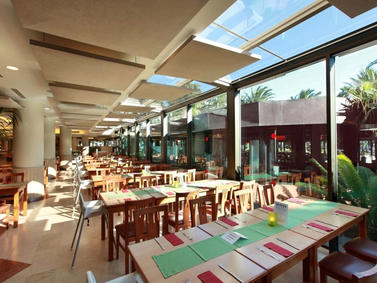 A long buffet restaurant with wooden tables laid with green and pink mats, under a partly glazed roof, with full-height windows onto palms and gardens.