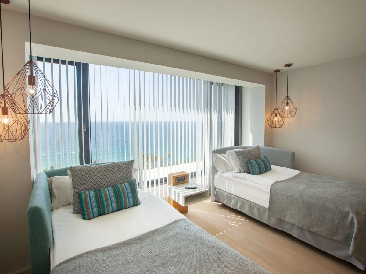 A room with two single beds under copper wire pendant lights, a wooden floor and a full-height window with white vertical blinds onto the sea.