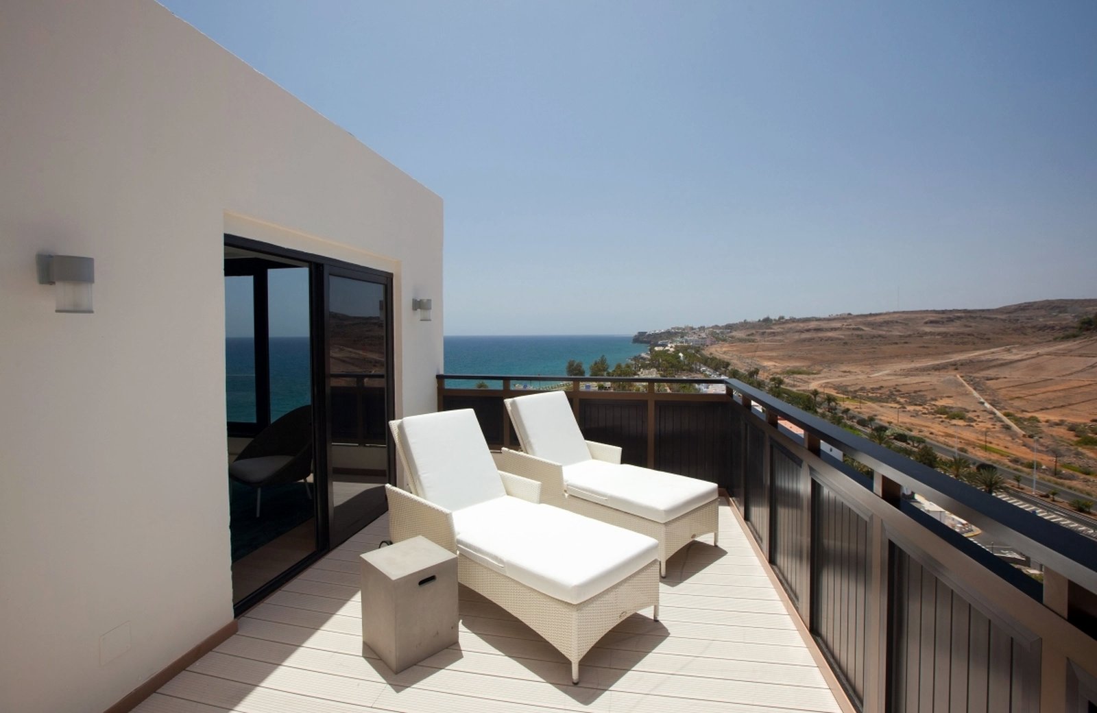 A wide terrace with two white sun loungers beside sliding doors, above the sea and the dry hillsides of Gran Canaria's south coast.