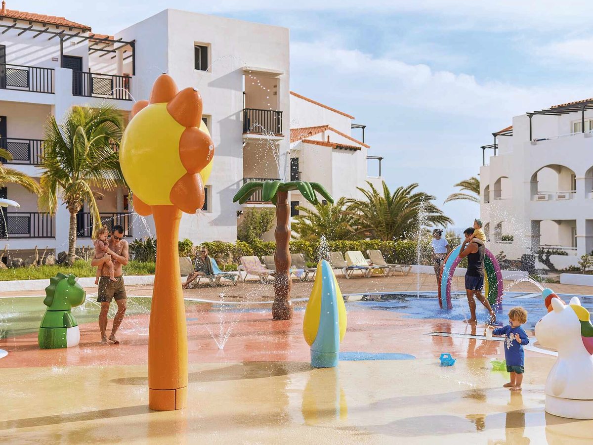 Children playing in a shallow splash area with brightly coloured water-play features and sprays, with white apartment blocks and palms behind.