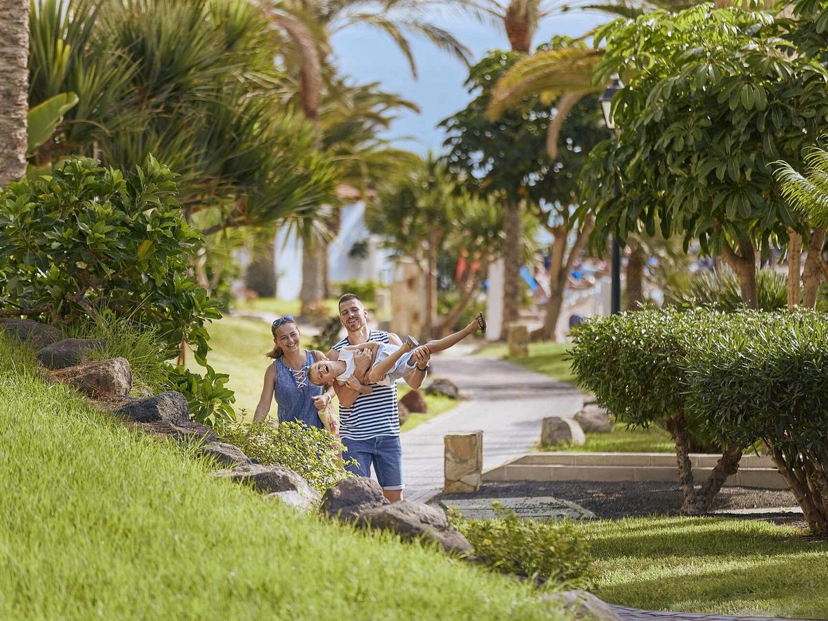 A family walking along a paved path through the resort gardens, past clipped lawns, palms and shrubs, with white accommodation blocks beyond.