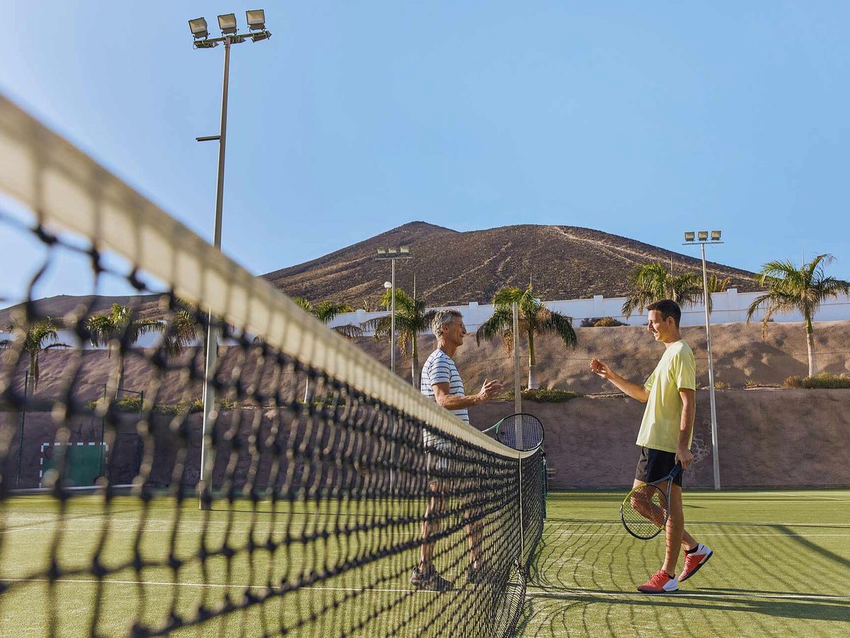 Two players on a green outdoor tennis court seen from behind the net, with floodlight poles, palms and a brown volcanic hill behind.