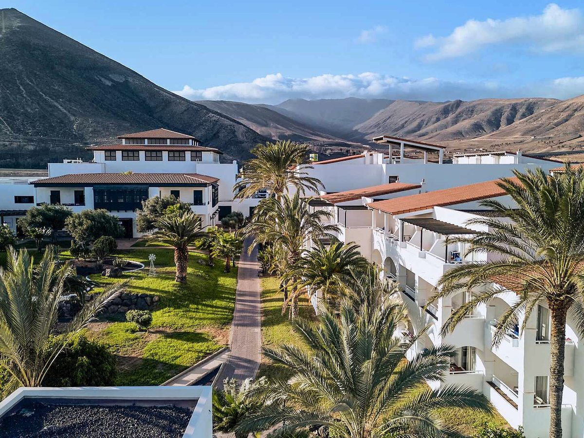 Landscaped resort gardens with palms and lawns in front of white two-storey blocks with arched balconies, and brown volcanic hills rising behind under a blue sky.